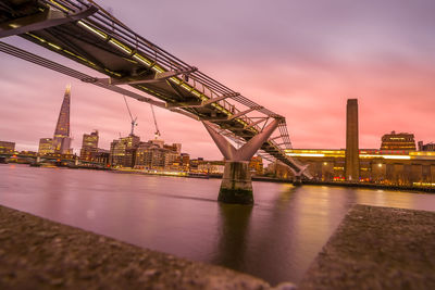 Illuminated bridge over river by buildings against sky at sunset