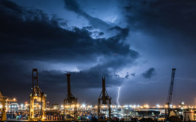 Cranes at harbor against cloudy sky at night