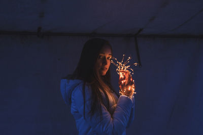 Beautiful young woman holding illuminated string light against wall