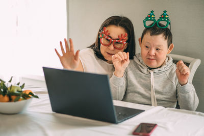 Girl helps elderly woman with down syndrome in funny christmas glasses, video call from laptop