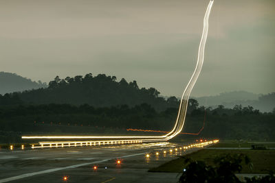 Light trails on road by trees against sky at night