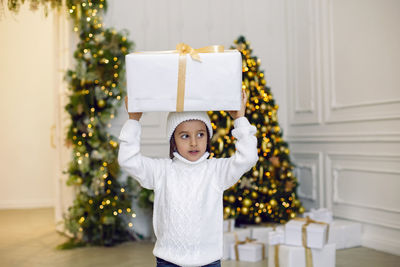 Boy in a white knitted sweater and hat stands with gift box  christmas tree at home on christmas day