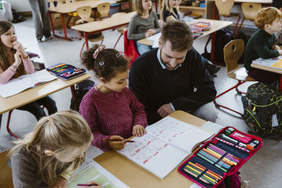 High angle view of male teacher crouching by schoolgirl solving mathematics in classroom