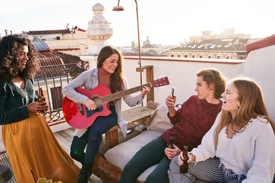 Cheerful young multiracial friends playing acoustic guitar and recording video on smartphone while having party on sunny rooftop