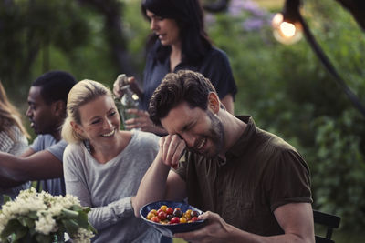 Happy woman looking at man rubbing eyes while holding tomato bowl at dinner party in yard