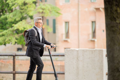 Side view full length young businessman in a suit riding an electric scooter while commuting to work 