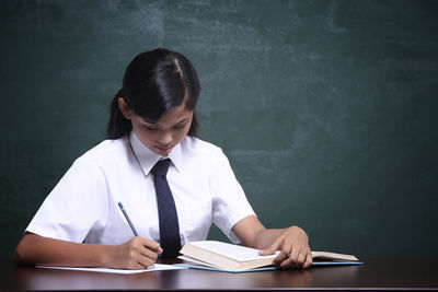 Schoolgirl studying at table against blackboard