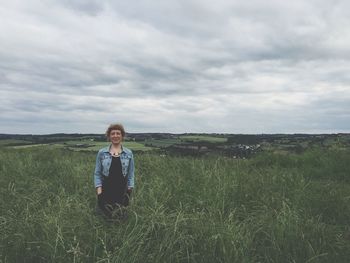 Smiling woman standing amidst grass on field against cloudy sky