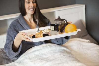 Woman brunette holding breakfast tray with tea cup,cake and fruits in bed at home in the bedroom