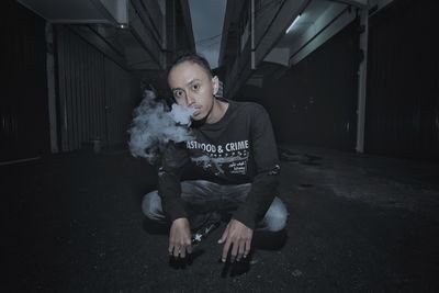 Portrait of young man smoking while crouching on street amidst buildings in city at night