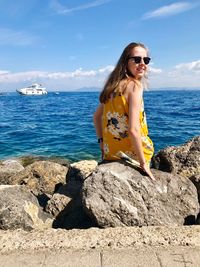 Portrait of smiling young woman sitting on rocks by sea against sky