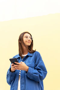 From below of young female in trendy outfit messaging on mobile phone on background of wall in city street and looking away