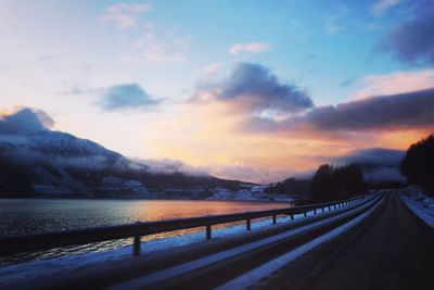 Country road by river against sky during sunset