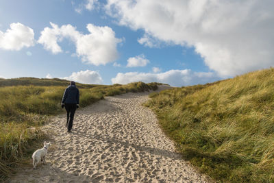 Rear view of woman and dog walking on sand dune against cloudy sky