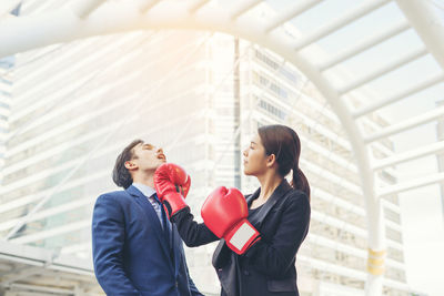 Young businesswoman hitting colleague with red boxing gloves against buildings in city