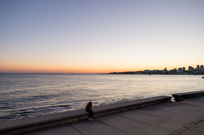 Woman sitting on retaining wall at promenade by sea against sky during sunset