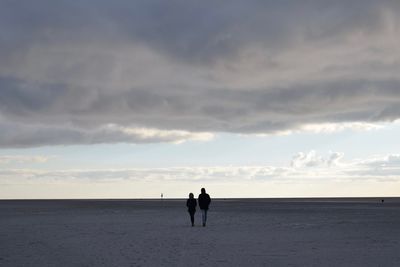 Rear view of friends walking at beach against cloudy sky