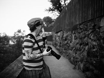 Side view of man holding camera while leaning on railing at footpath