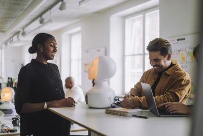 Multiracial smiling students with illuminated social robot at desk in innovation lab