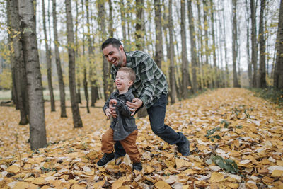 Cheerful father and son playing on dry leaves in forest during autumn