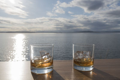 Whiskey glasses on table by sea against sky