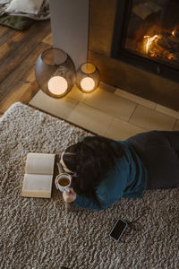 High angle view of woman reading book while lying on carpet at home