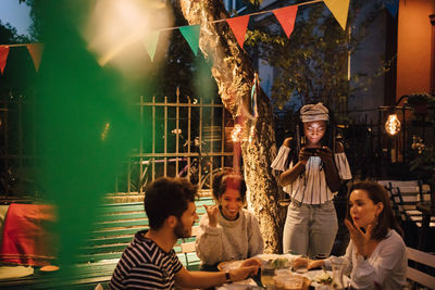 Young woman using mobile phone while friends talking at table during dinner party