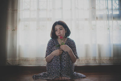 Full length of thoughtful woman holding rose while sitting against window