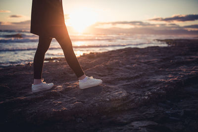 Low section of woman standing on beach against sky during sunset