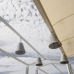 Low angle view of lighting equipment hanging on ceiling against cloudy sky