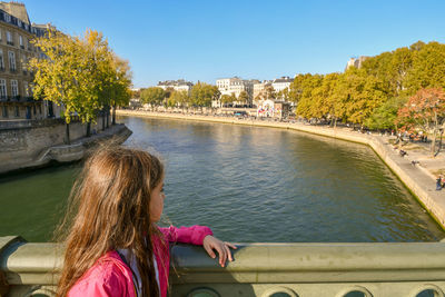 Side view of girl standing by railing while looking away by river against sky