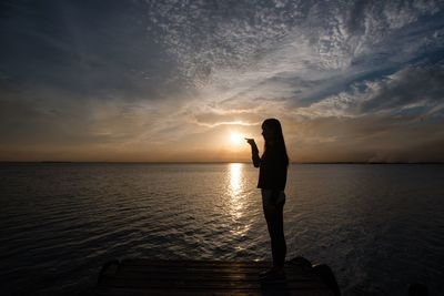 Optical illusion of woman pointing sun while standing on jetty by lake during sunset