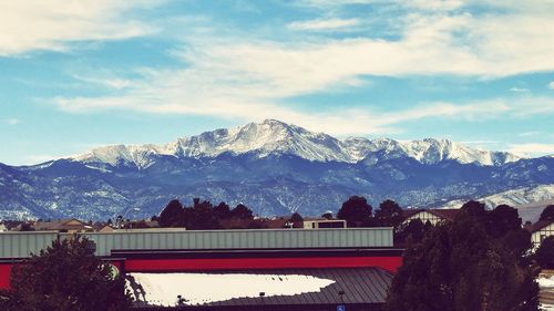 Scenic view of mountains in front of buildings against cloudy sky during winter