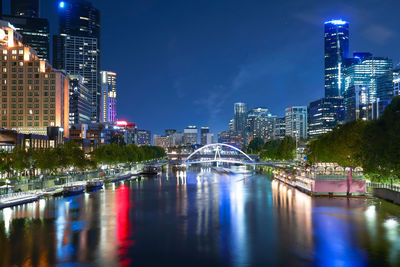 Illuminated bridge over river by buildings against sky at night