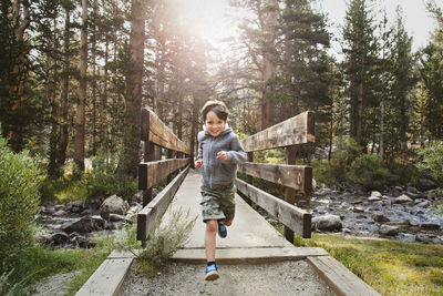 Portrait of boy running over wooden bridge against trees in forest