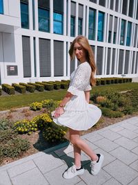 Smiling young woman in white dress walking against building