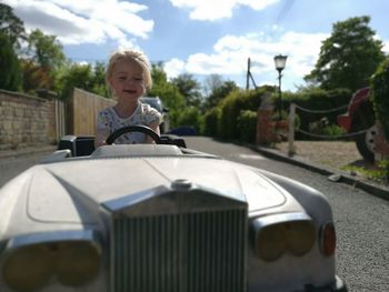 Smiling girl driving toy car on road by trees against sky