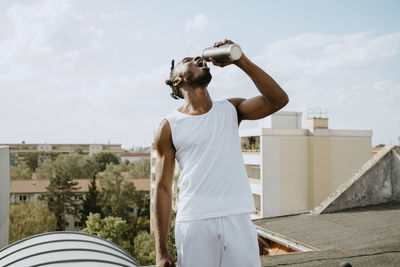 Young man drinking water from bottle while standing on rooftop during sunny day