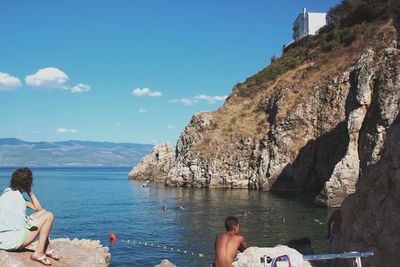 People relaxing on rock by sea against sky