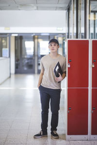 Portrait of confident teenage boy holding book while standing by lockers in corridor at high school