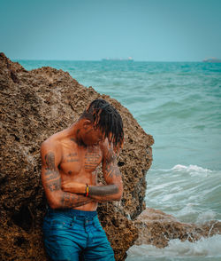 Midsection of shirtless man on rock at beach against sky
