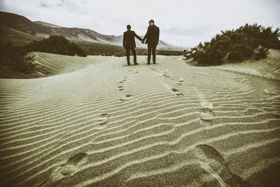 Rear view of gay men holding hands while standing on sand at desert against sky