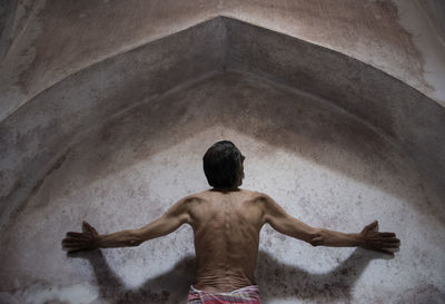 Rear view of shirtless man with arms outstretched against wall