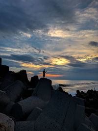 Man standing on rock at beach against sky during sunset