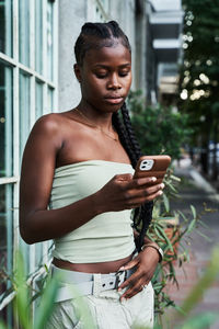 Young african american woman with long braids wearing stylish tube top and browsing smartphone on city street