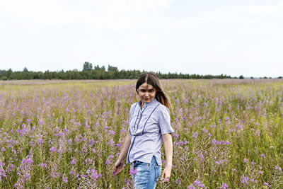 Young beautiful smiling blond woman in purple shirt walking in meadow among flowers fireweed,  field