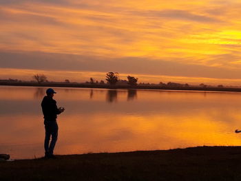 Silhouette man standing by lake against sky during sunset