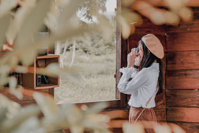 Side view of woman photographing while standing by window