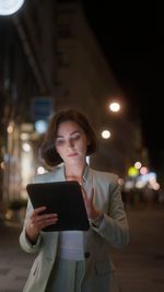 Portrait of young woman using mobile phone while standing in city at night