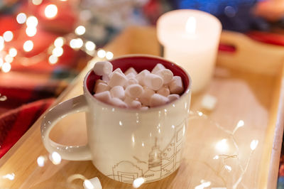 Cup of cocoa and marshmallow and burning candle on tray on red plaid and a garland of glowing lights